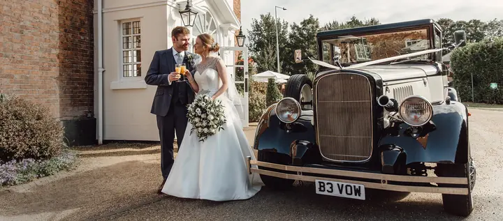 A couple raising a glass beside a vintage wedding car