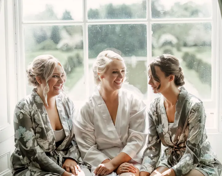 A bride and her bridesmaids laughing together in matching robes by a window