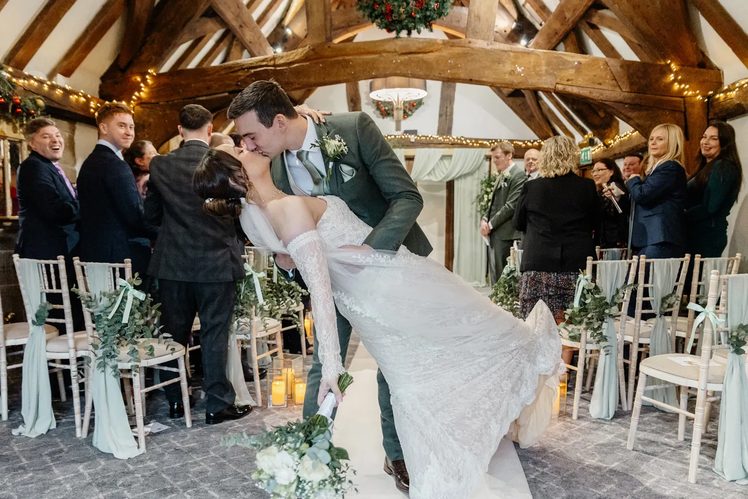 Groom dipping his bride for a kiss in a candlelit barn as guests cheer