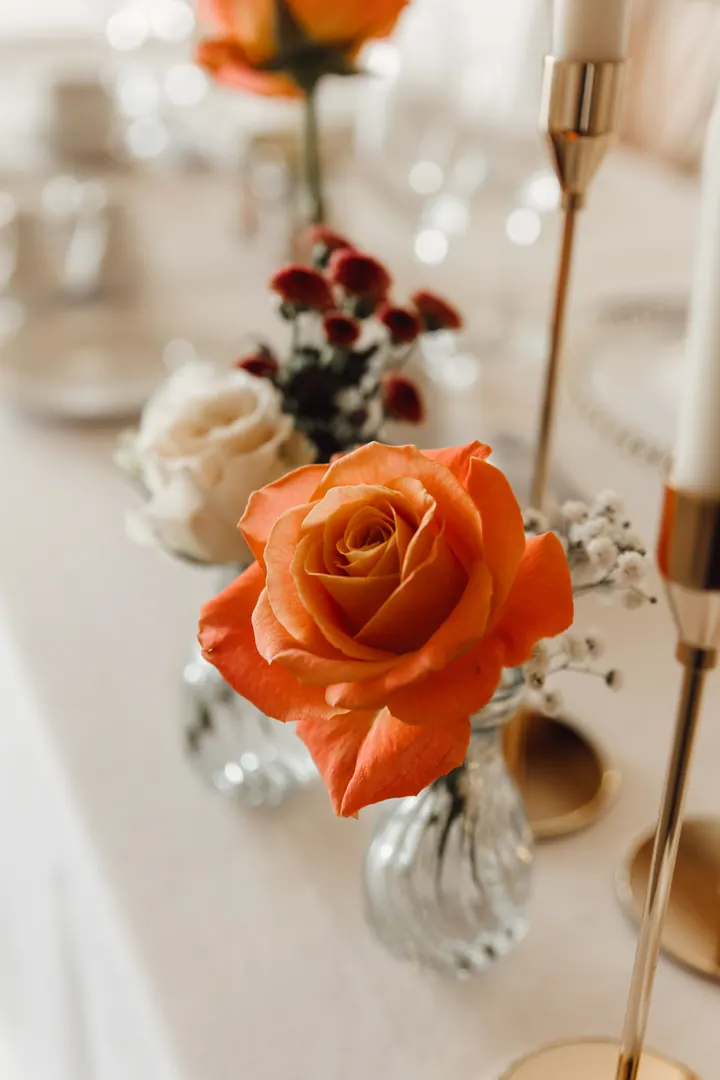 A single orange rose in a bud vase beside gold candlesticks