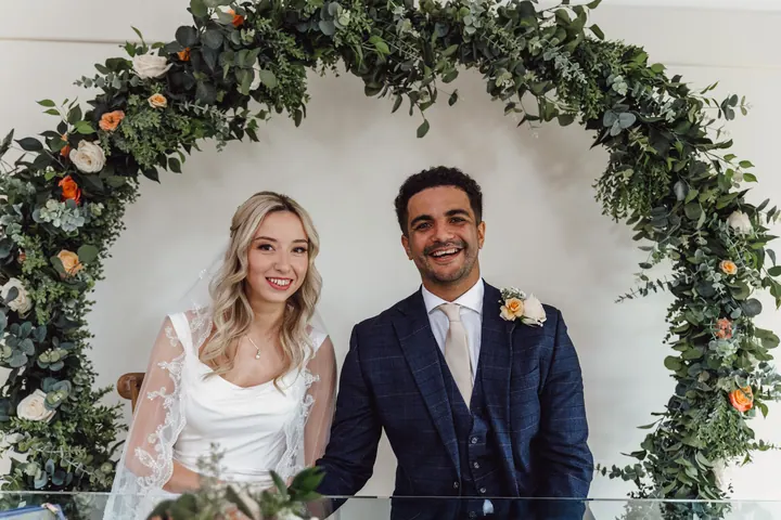 The couple laughing as they sign the register beneath a hoop of greenery and roses