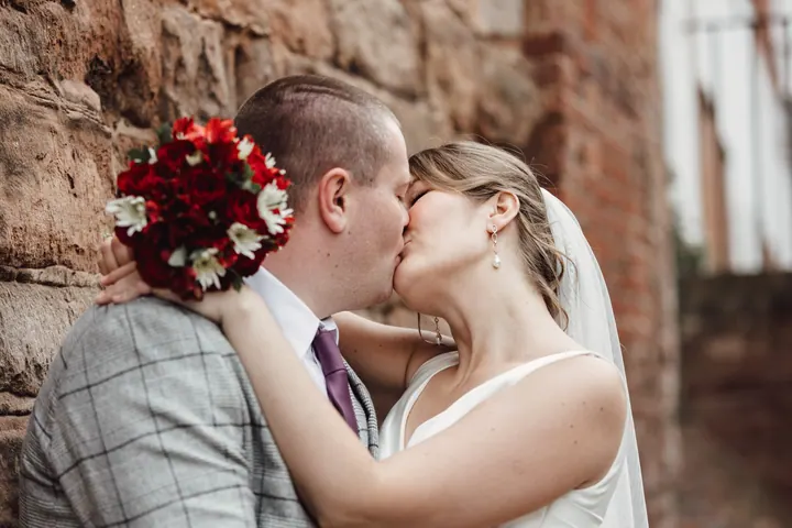 A kiss against the sandstone wall, bouquet over Jamie&rsquo;s shoulder