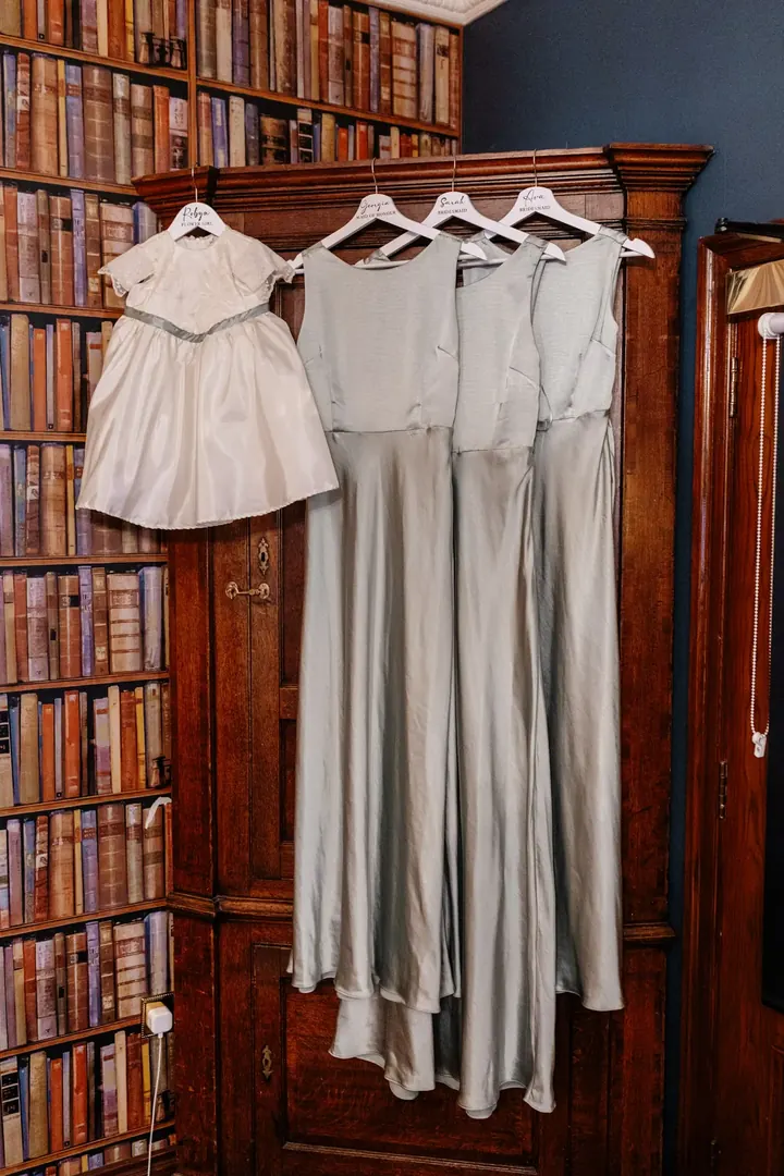Sage green bridesmaid dresses hanging from an old wooden bookcase