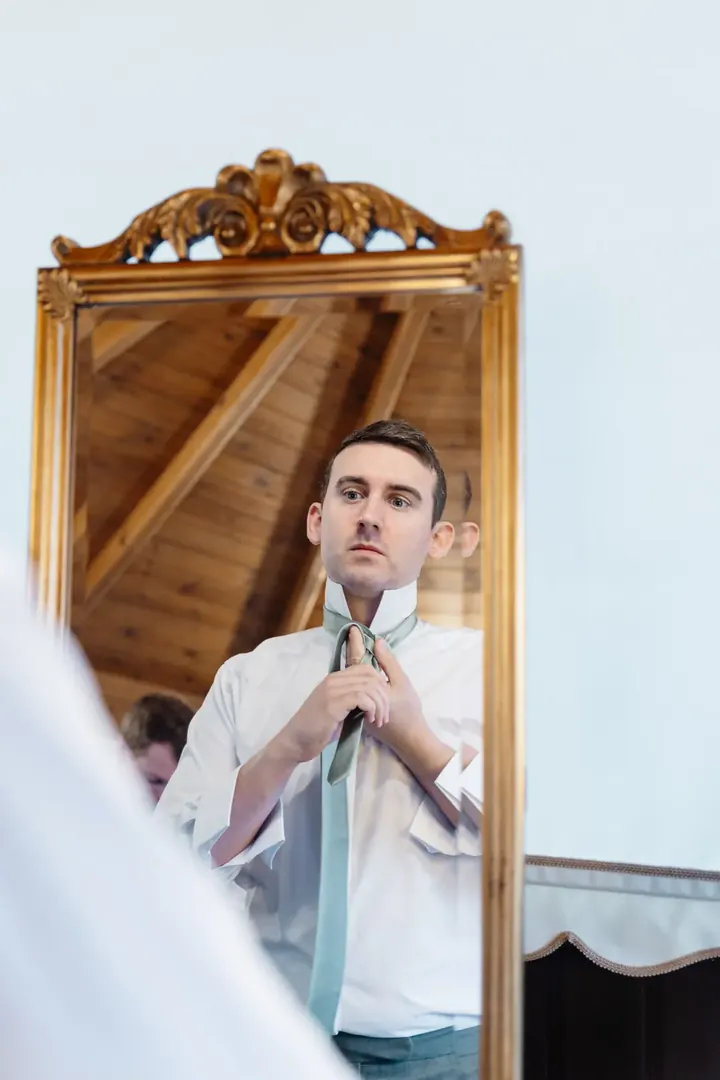 Andrew fastening his sage tie in a gilded mirror