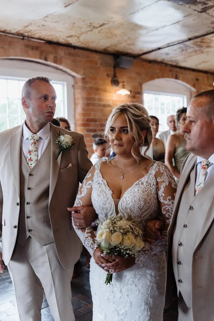 A bride walking down the aisle between two smiling men in beige suits