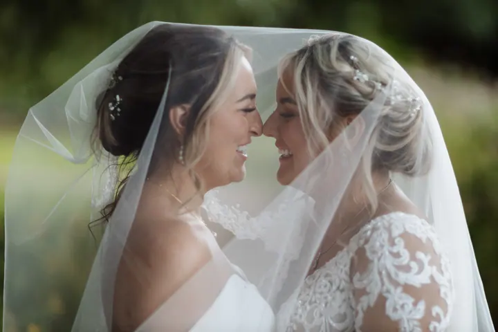 Two brides nose to nose beneath a shared veil at The West Mill